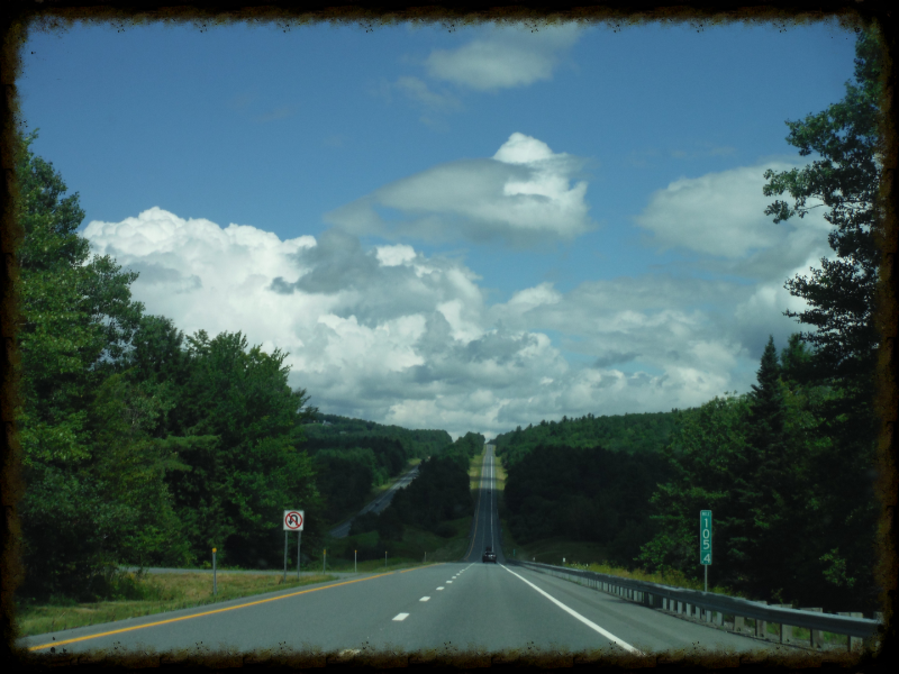 A long, straight road stretches under a partly cloudy sky through a green, forested landscape.